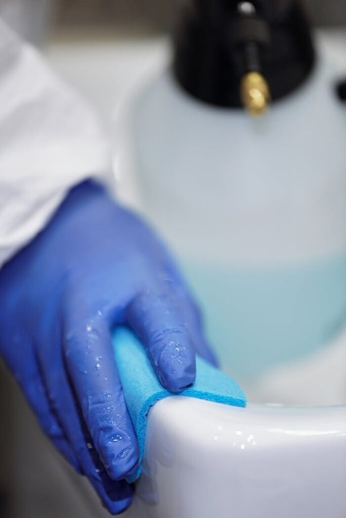 A blue-gloved hand using a sponge to clean a white sink in a hygienic environment.